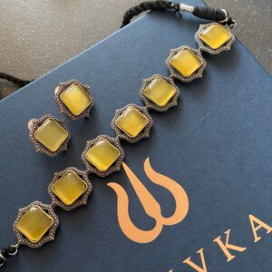 May include: A set of yellow gemstone earrings and a necklace with a silver finish. The necklace has a black cord and the earrings are stud style. The jewelry is displayed on a blue box with the word "SHIVKA" in gold lettering.