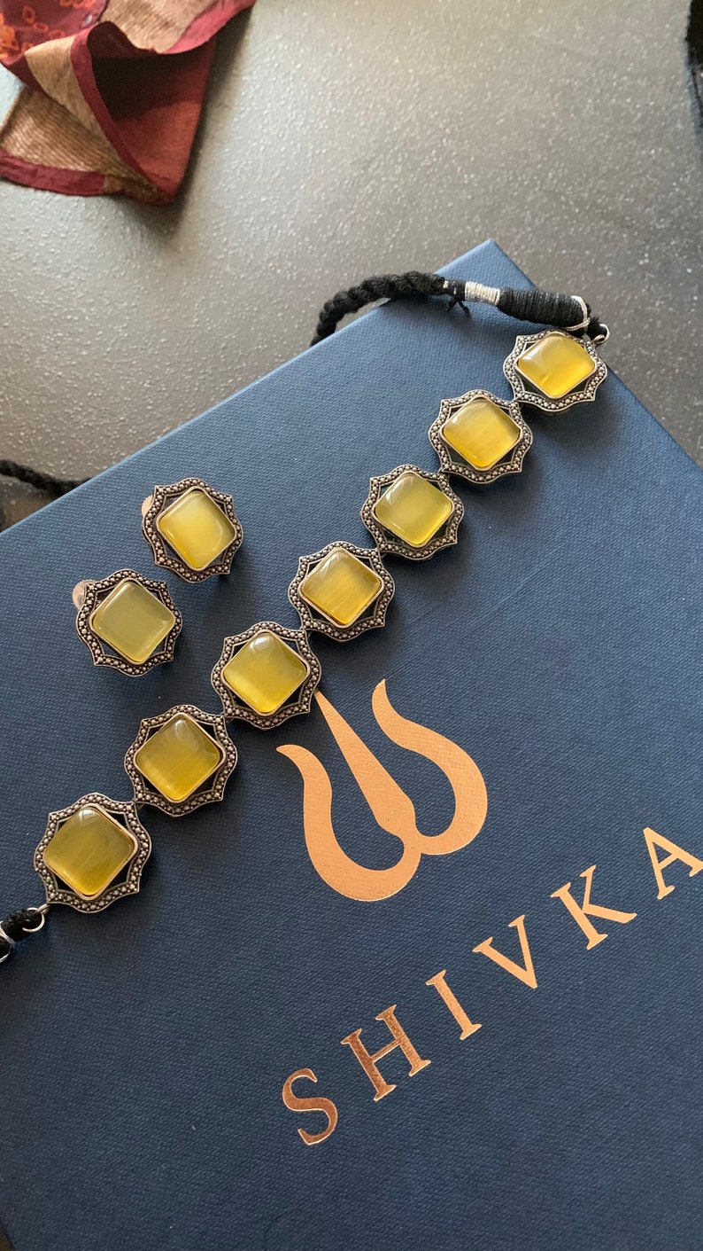 May include: A set of yellow gemstone earrings and a necklace with a silver finish. The necklace has a black cord and the earrings are stud style. The jewelry is displayed on a blue box with the word "SHIVKA" in gold lettering.