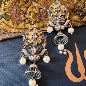 May include: A pair of silver earrings with a floral design and pearl accents. The earrings feature a round, yellow-toned center with a clear stone. The earrings are hanging from a silver chain with a bell-shaped charm.