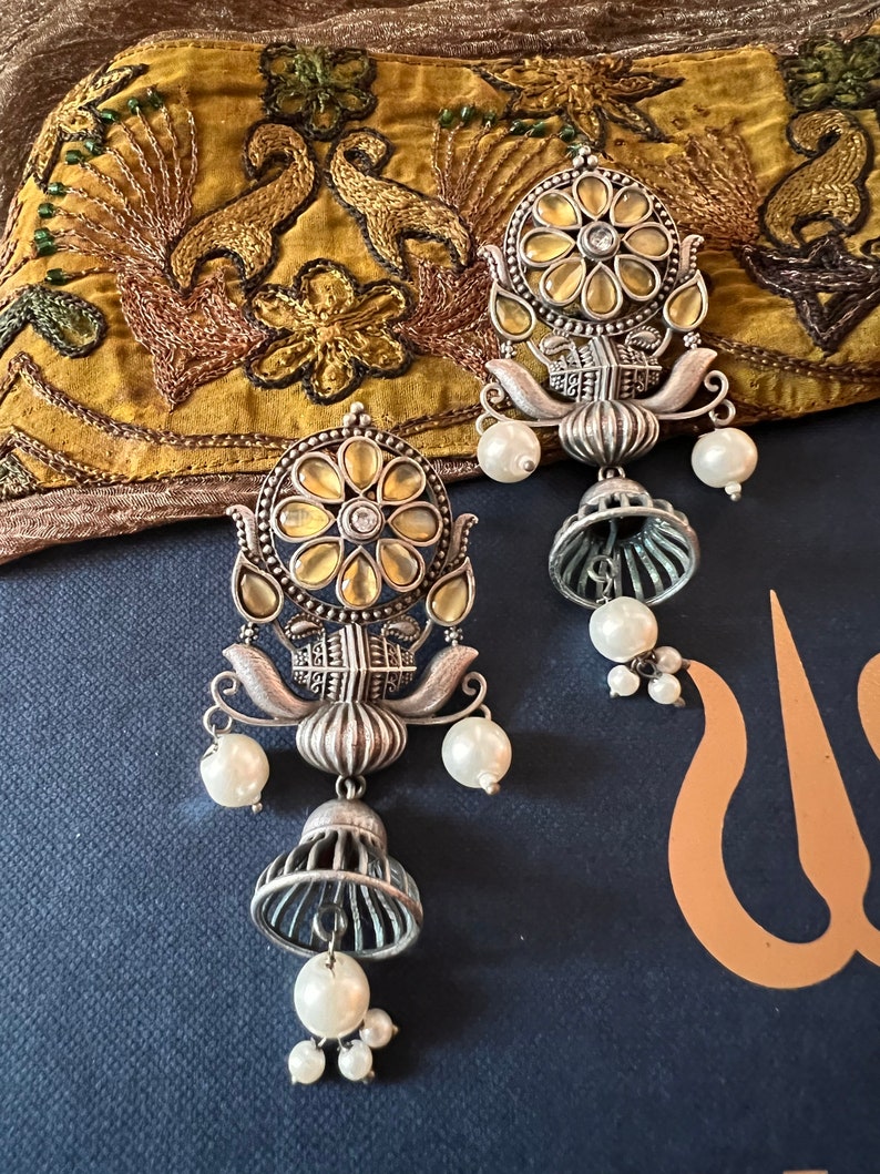 May include: A pair of silver earrings with a floral design and pearl accents. The earrings feature a round, yellow-toned center with a clear stone. The earrings are hanging from a silver chain with a bell-shaped charm.