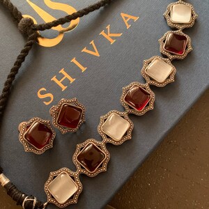 May include: A silver necklace with red and white square-shaped stones set in ornate silver frames. The necklace is on a blue box with the word "SHIVIKA" printed on it.