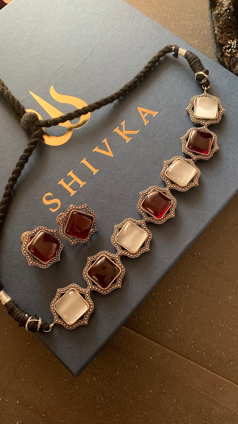 May include: A silver necklace with red and white square-shaped stones set in ornate silver frames. The necklace is on a blue box with the word "SHIVIKA" printed on it.