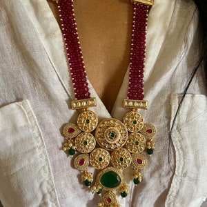May include: A long gold necklace with a large pendant featuring intricate details and green and red gemstones. The necklace is made of red beads and has a gold chain with rectangular accents.