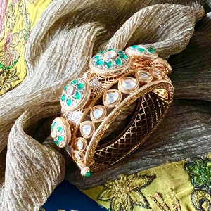 K&ouml;nnte beinhalten: Zwei goldene Armreifen mit filigranen Designs und gr&uuml;nen und wei&szlig;en Edelsteinen. Die Armreifen liegen auf einem gelben und gr&uuml;nen Stoff mit dem Wort "SHIV" auf einer blauen Oberfl&auml;che.