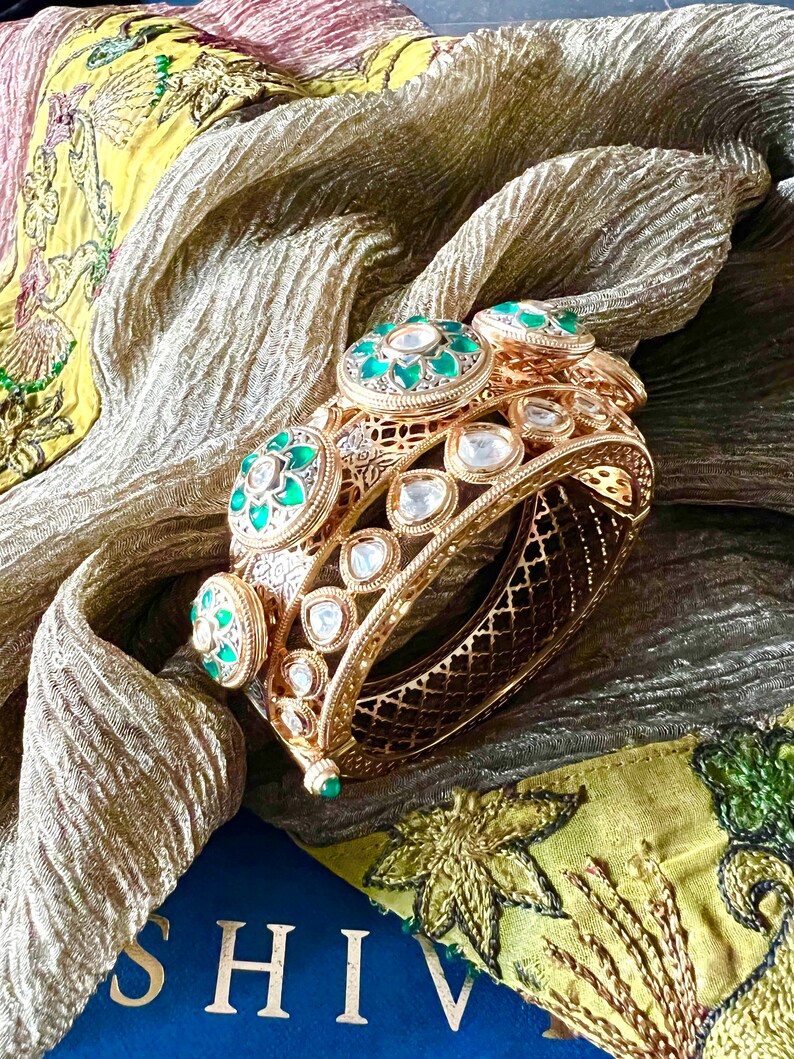 K&ouml;nnte beinhalten: Zwei goldene Armreifen mit filigranen Designs und gr&uuml;nen und wei&szlig;en Edelsteinen. Die Armreifen liegen auf einem gelben und gr&uuml;nen Stoff mit dem Wort "SHIV" auf einer blauen Oberfl&auml;che.