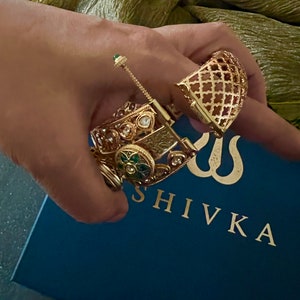 K&ouml;nnte beinhalten: Zwei goldene Armreifen mit aufwendigen Designs und bunten Edelsteinen. Die Armreifen liegen auf einer blauen Box mit dem in Gold gedruckten Wort "SHIVKA".
