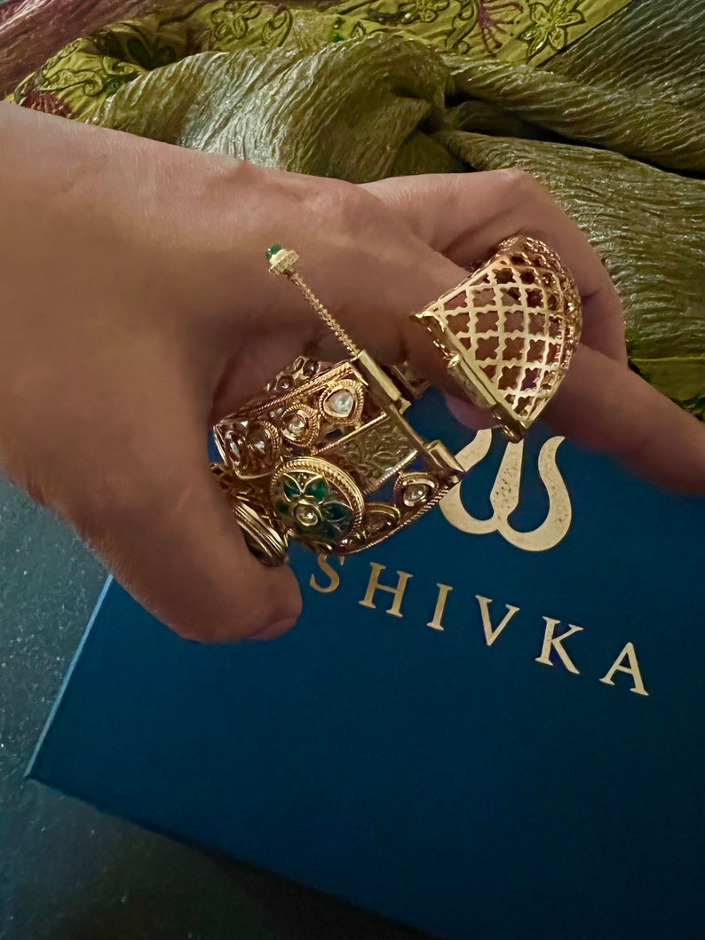 K&ouml;nnte beinhalten: Zwei goldene Armreifen mit aufwendigen Designs und bunten Edelsteinen. Die Armreifen liegen auf einer blauen Box mit dem in Gold gedruckten Wort "SHIVKA".