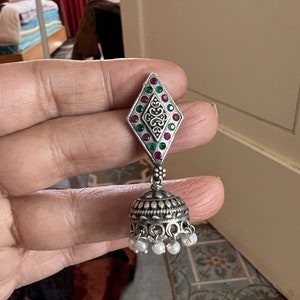May include: Silver-toned earring with a diamond-shaped top adorned with red and green gemstones and a central filigree design. The bottom features a bell-shaped element with small white pearl accents, creating a traditional, ornate look.