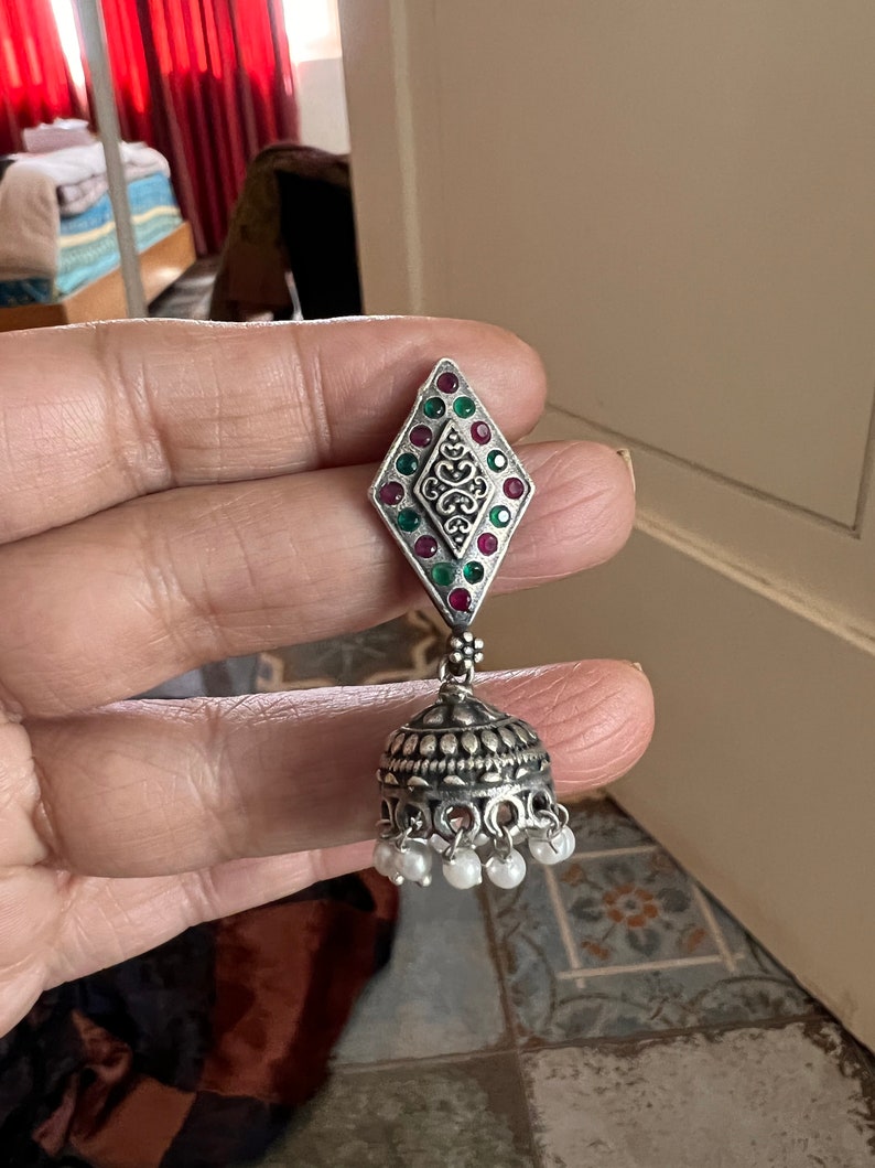 May include: Silver-toned earring with a diamond-shaped top adorned with red and green gemstones and a central filigree design. The bottom features a bell-shaped element with small white pearl accents, creating a traditional, ornate look.