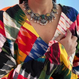 May include: A silver-toned statement necklace with black, yellow, and green gemstones and small white pearls. The necklace is worn over a colorful patchwork patterned top with a V-neck.