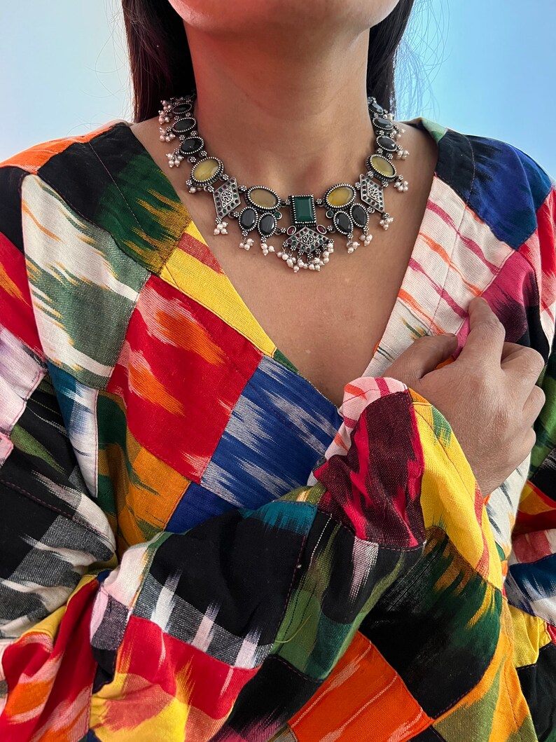 May include: A silver-toned statement necklace with black, yellow, and green gemstones and small white pearls. The necklace is worn over a colorful patchwork patterned top with a V-neck.