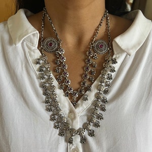 May include: Silver-toned necklace with a layered design. The necklace features a floral pattern with small red gemstones and dangling bell-shaped charms. The necklace is worn over a white collared shirt.