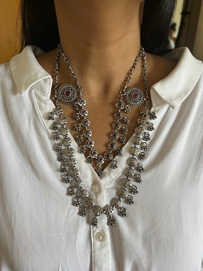 May include: Silver-toned necklace with a layered design. The necklace features a floral pattern with small red gemstones and dangling bell-shaped charms. The necklace is worn over a white collared shirt.