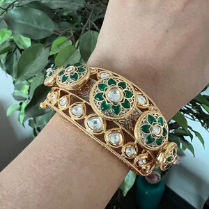 K&ouml;nnte beinhalten: Ein goldenes Armreif-Armband mit gr&uuml;nem und wei&szlig;em Emaille und klaren Steinen. Das Armband ist mit floralen Designs verziert.