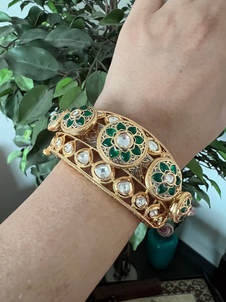 K&ouml;nnte beinhalten: Ein goldenes Armreif-Armband mit gr&uuml;nem und wei&szlig;em Emaille und klaren Steinen. Das Armband ist mit floralen Designs verziert.