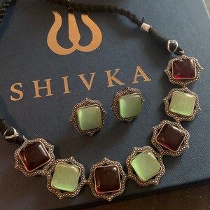 May include: A silver necklace with a black cord and a pendant featuring alternating green and red square stones. The necklace is displayed on a blue box with the word "SHIVKA" printed on it.