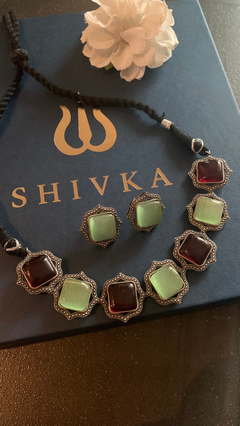 May include: A silver necklace with a black cord and a pendant featuring alternating green and red square stones. The necklace is displayed on a blue box with the word "SHIVKA" printed on it.