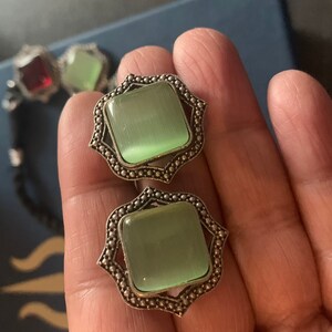 May include: A pair of silver earrings with green cat's eye stones set in a square shape with a decorative border.