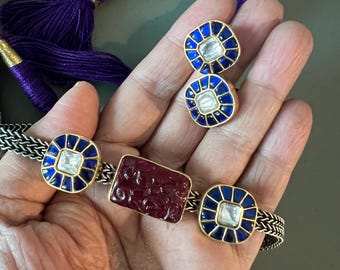 May include: A gold necklace with a red stone and blue enamel accents. The necklace is on a chain with a tassel. There are also two matching earrings with blue enamel and a clear stone.