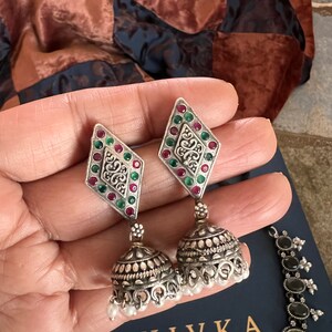 May include: Silver-toned earrings with a diamond-shaped top adorned with red and green gemstones, and a bell-shaped bottom with small white pearls. The earrings are part of a jewelry set, with the brand name "SHIVKA" visible.