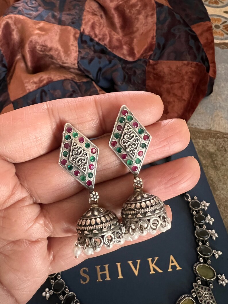 May include: Silver-toned earrings with a diamond-shaped top adorned with red and green gemstones, and a bell-shaped bottom with small white pearls. The earrings are part of a jewelry set, with the brand name "SHIVKA" visible.