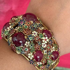 May include: A wide gold cuff bracelet featuring large, smooth, deep red cabochon stones interspersed with an intricate openwork pattern of small, multi-colored enamel flowers in shades of white, blue, green, and red, with a hinged closure.