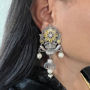 May include: Silver earrings with a floral design and yellow and white stones. The earrings have a dangling bell shape with pearl accents.