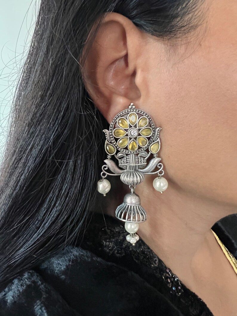 May include: Silver earrings with a floral design and yellow and white stones. The earrings have a dangling bell shape with pearl accents.