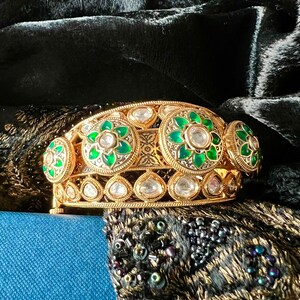 K&ouml;nnte beinhalten: Ein goldenes Armreif-Armband mit gr&uuml;nen und wei&szlig;en Edelsteinen. Das Armband ist mit komplizierten floralen Designs verziert und vor einem schwarzen Samt-Hintergrund platziert.