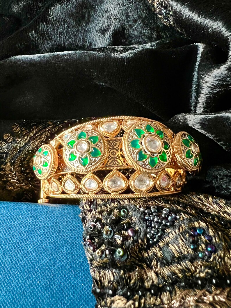 K&ouml;nnte beinhalten: Ein goldenes Armreif-Armband mit gr&uuml;nen und wei&szlig;en Edelsteinen. Das Armband ist mit komplizierten floralen Designs verziert und vor einem schwarzen Samt-Hintergrund platziert.