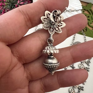 May include: A silver-toned earring with a floral stud and a dangling bell-shaped charm. The earring is part of a necklace, with a detailed design. The jewelry is displayed against a soft background, highlighting the intricate metalwork.