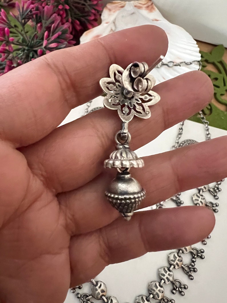 May include: A silver-toned earring with a floral stud and a dangling bell-shaped charm. The earring is part of a necklace, with a detailed design. The jewelry is displayed against a soft background, highlighting the intricate metalwork.