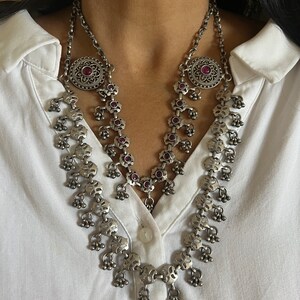 May include: Silver-toned necklace with a floral design and small dangling bells. The necklace features a large circular pendant with a red gemstone in the center. The necklace is worn over a white collared shirt.