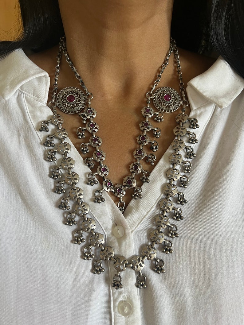 May include: Silver-toned necklace with a floral design and small dangling bells. The necklace features a large circular pendant with a red gemstone in the center. The necklace is worn over a white collared shirt.