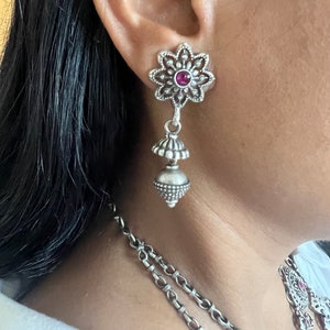 May include: Silver-toned dangle earrings with a floral design and a small, round, red gemstone in the center of each flower. The earrings have a tiered, bell-shaped drop. A matching necklace is partially visible.