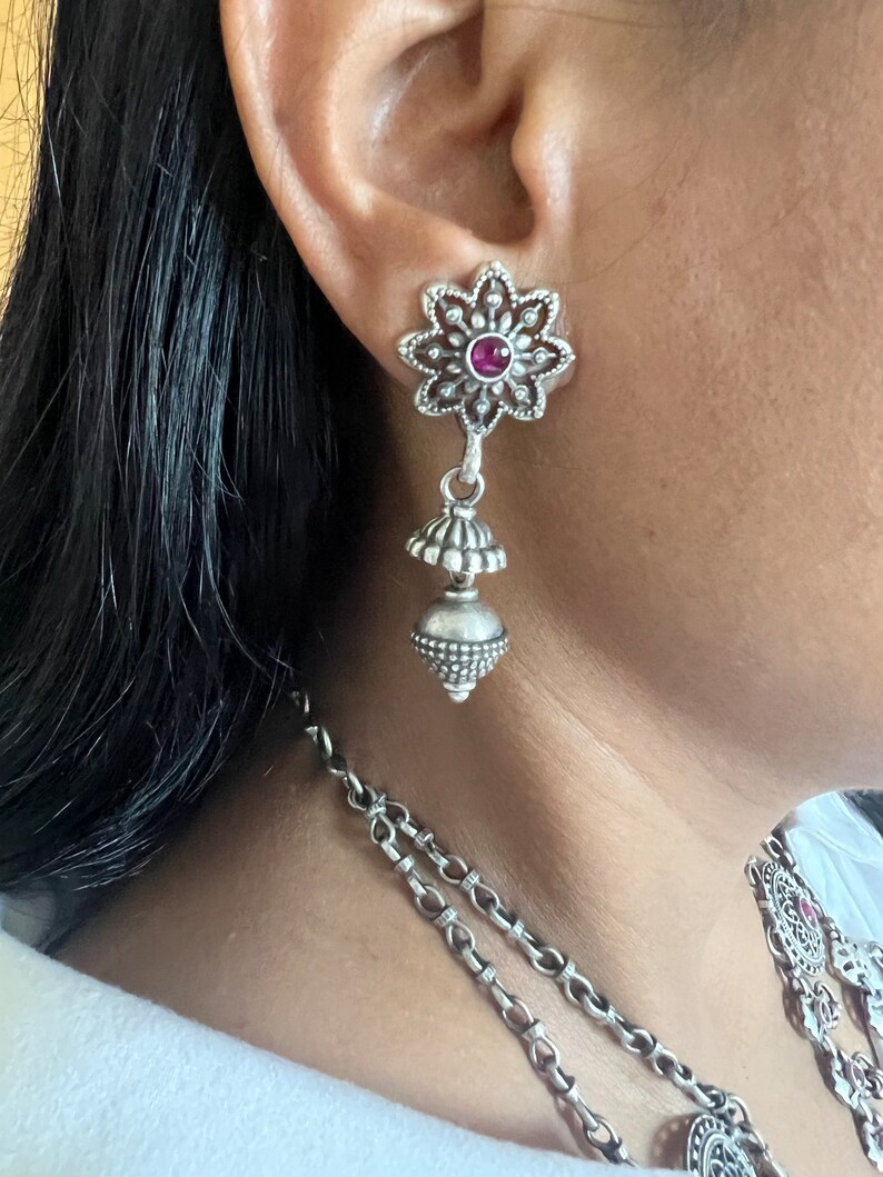 May include: Silver-toned dangle earrings with a floral design and a small, round, red gemstone in the center of each flower. The earrings have a tiered, bell-shaped drop. A matching necklace is partially visible.