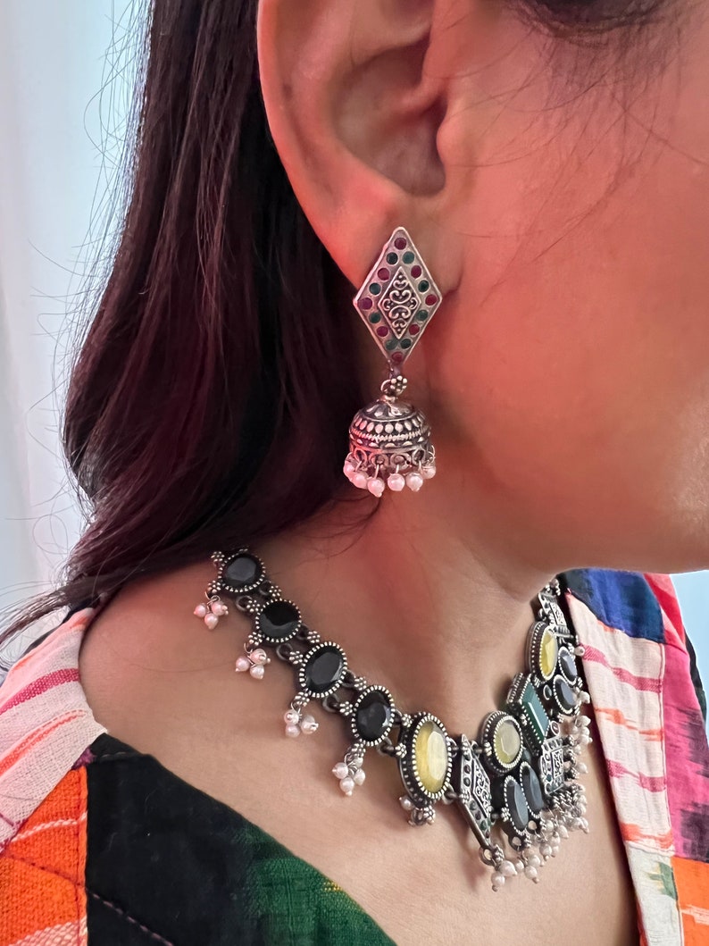 May include: Silver-toned necklace and earring set. The necklace features black and yellow oval stones, with small white pearl accents. The earrings have a diamond-shaped top with red, green, and silver accents, and a bell-shaped bottom with white pearl dangles.