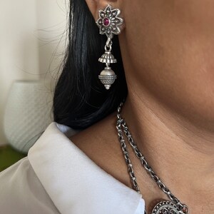 May include: Silver-toned dangle earrings with a floral design and a small red gemstone in the center. The earrings have a bell-shaped element and a beaded sphere. A matching necklace is also visible.