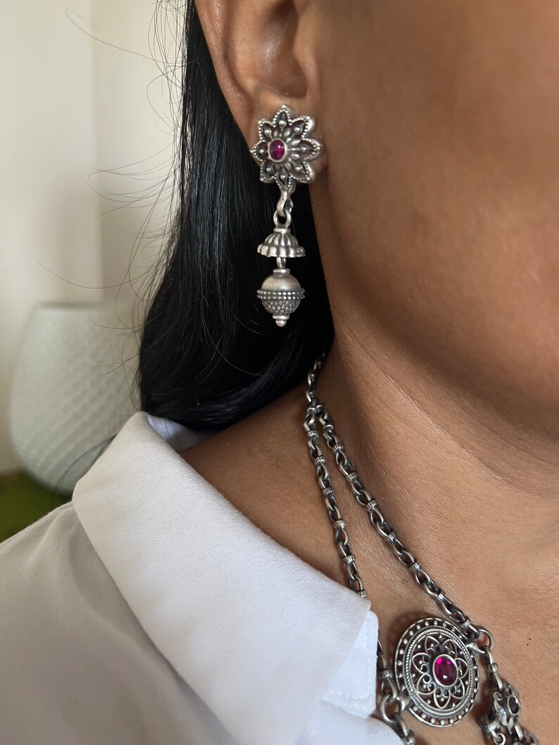 May include: Silver-toned dangle earrings with a floral design and a small red gemstone in the center. The earrings have a bell-shaped element and a beaded sphere. A matching necklace is also visible.