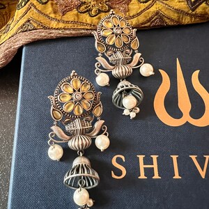 May include: A pair of silver earrings with a yellow and white floral design. The earrings have a bell-shaped dangle with a pearl at the bottom. The earrings are on a blue background with the word "SHIV" in gold lettering.