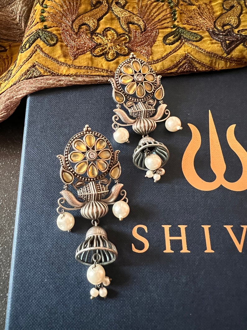 May include: A pair of silver earrings with a yellow and white floral design. The earrings have a bell-shaped dangle with a pearl at the bottom. The earrings are on a blue background with the word "SHIV" in gold lettering.