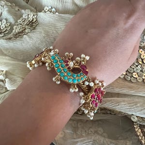 Op de afbeelding: Een gouden armband met turquoise en rode stenen, en parel accenten. De armband is in de vorm van een pauw.