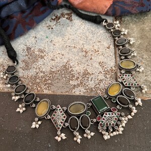 May include: A silver-toned necklace featuring a variety of gemstones in oval, square, and diamond shapes. The stones are in shades of black, yellow, and green, with small white pearl accents. The necklace has a black cord.
