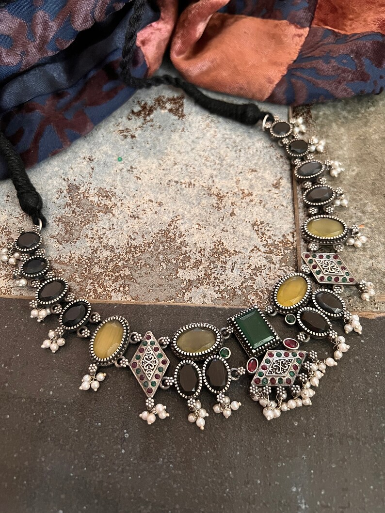 May include: A silver-toned necklace featuring a variety of gemstones in oval, square, and diamond shapes. The stones are in shades of black, yellow, and green, with small white pearl accents. The necklace has a black cord.
