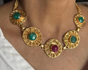 Puede incluir: Collar de oro con cinco colgantes redondos. Cada colgante presenta un diseño tejido y una piedra preciosa en el centro. Las piedras preciosas son verdes y rojas.