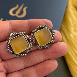 May include: A pair of silver earrings with a yellow gemstone in the center of each earring. The earrings have a decorative, ornate design.