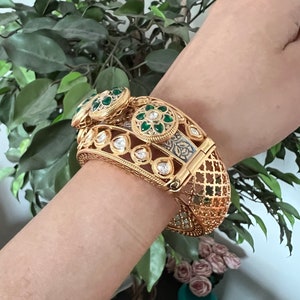 K&ouml;nnte beinhalten: Ein goldenes Armreif-Armband mit aufwendigen Details und gr&uuml;nen und wei&szlig;en Edelsteinen. Das Armband ist aus Metall und hat ein filigranes Design.