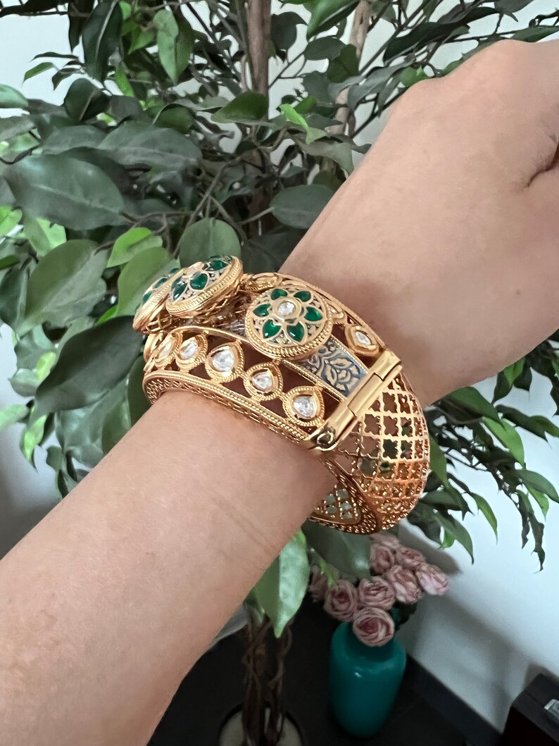 K&ouml;nnte beinhalten: Ein goldenes Armreif-Armband mit aufwendigen Details und gr&uuml;nen und wei&szlig;en Edelsteinen. Das Armband ist aus Metall und hat ein filigranes Design.