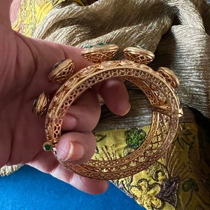 K&ouml;nnte beinhalten: Ein goldenes Armreif-Armband mit filigranen Details und gr&uuml;nen Edelstein-Akzenten.