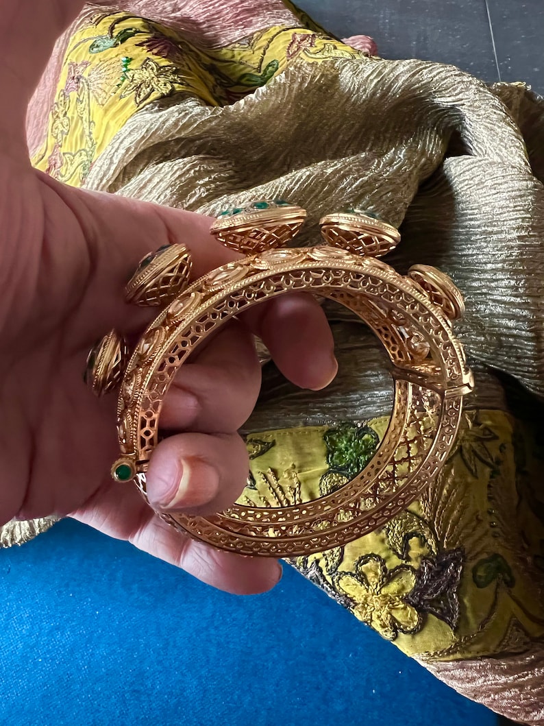 K&ouml;nnte beinhalten: Ein goldenes Armreif-Armband mit filigranen Details und gr&uuml;nen Edelstein-Akzenten.